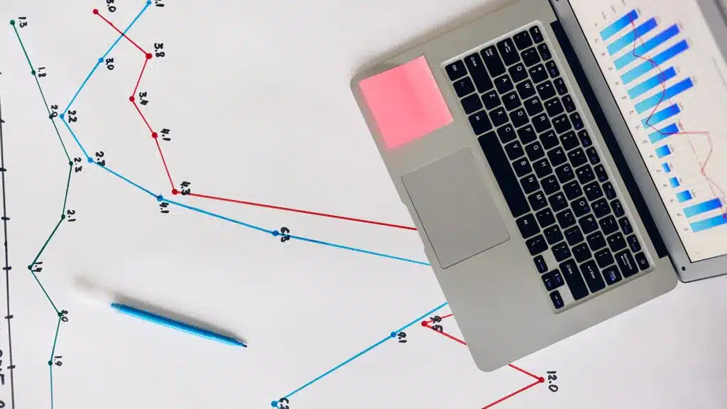 A laptop displaying graphs and charts on a desk with a blue pen and handwritten data points in the background.